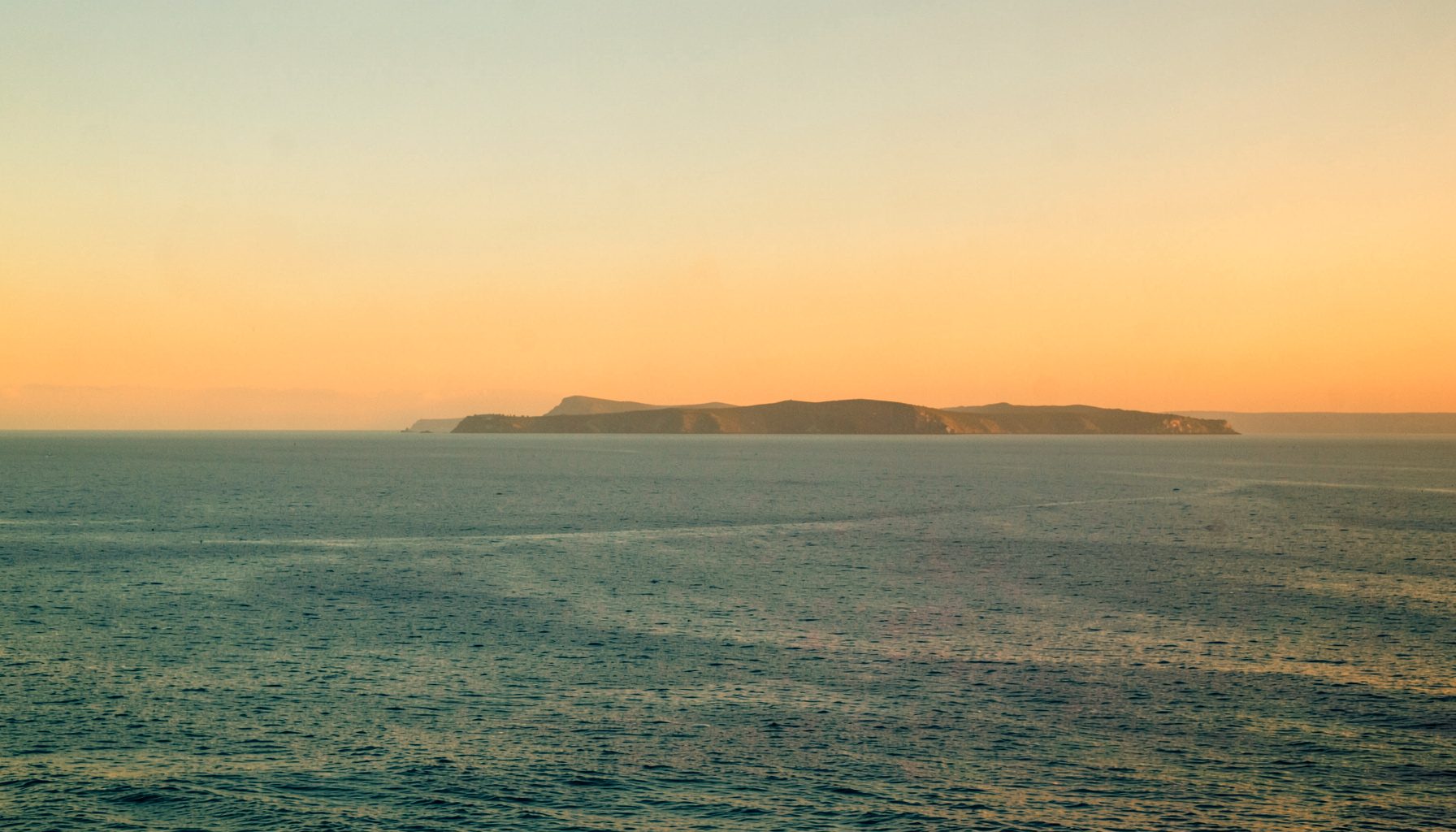 Wide coastal horizon at dawn, soft amber light catching on calm water and a low band of distant headland, rendered in muted blues and warm golds.